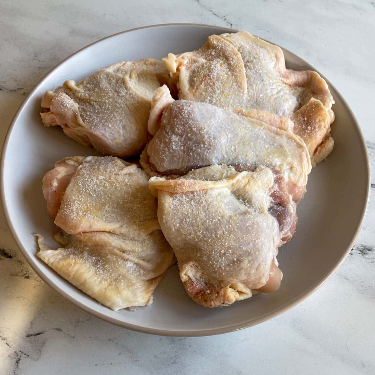 Raw chicken thighs dusted with kosher salt sit on a white plate.