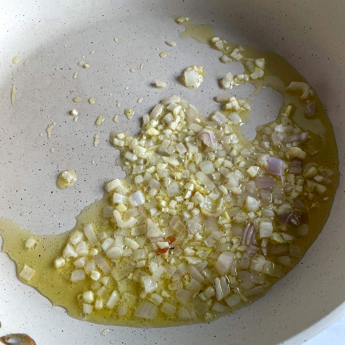 Garlic and shallot are shown sweating in olive oil in a white frying pan.