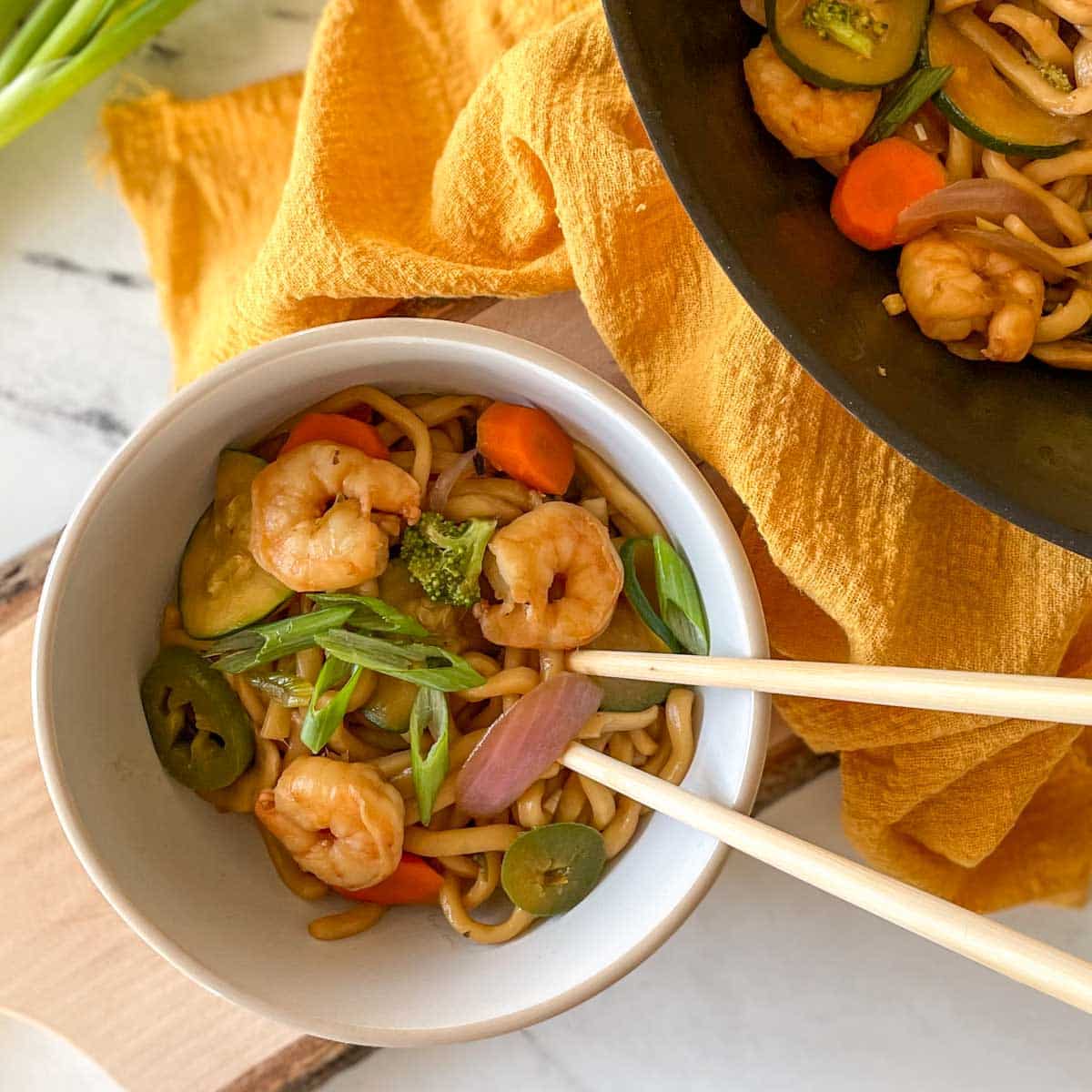 A white bowl of Shrimp Yaki Udon sits on a rustic wooden cutting board with chopsticks with a yellow linen and green onions.