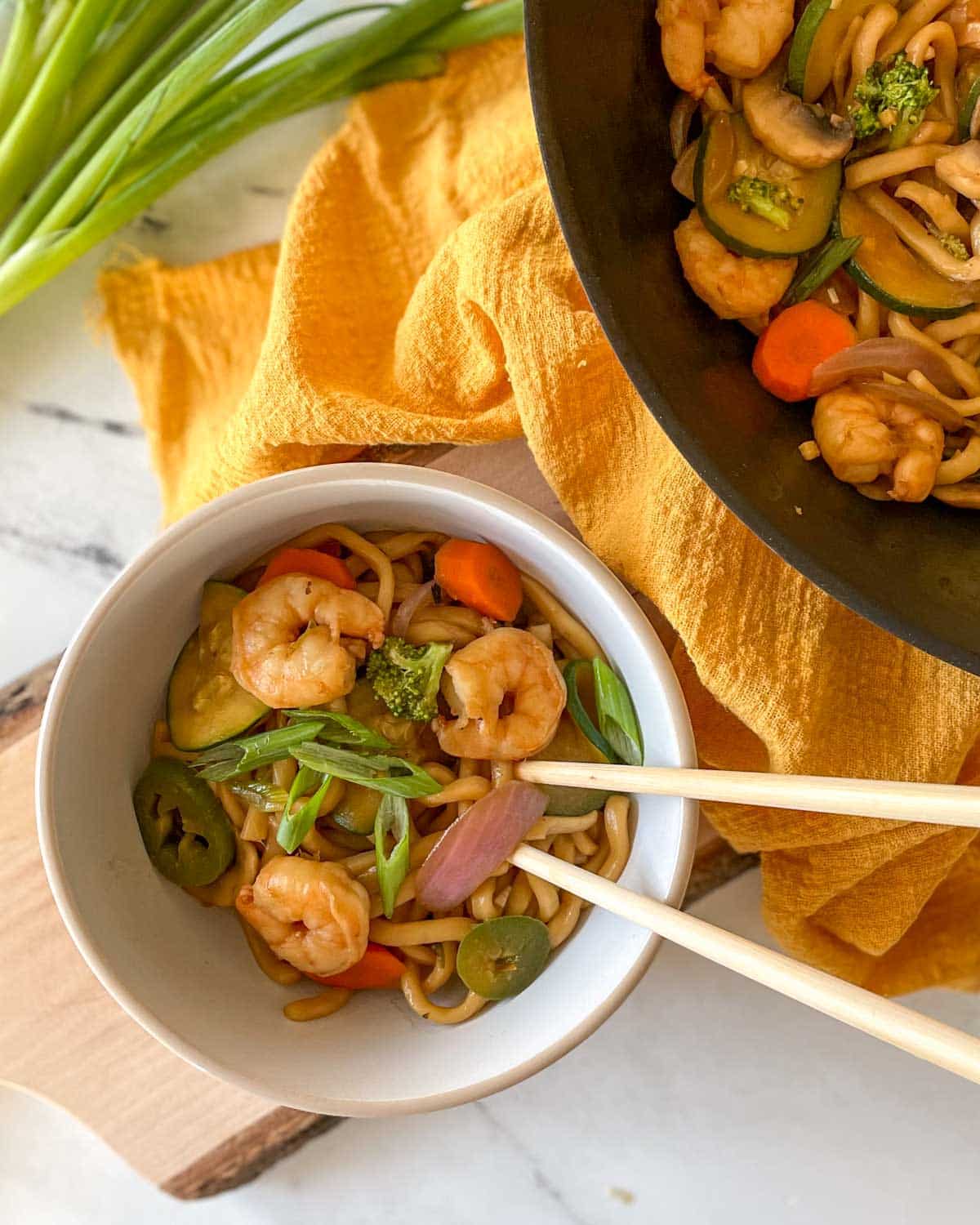 A white bowl of Shrimp Yaki Udon sits on a rustic wooden cutting board with chopsticks with a yellow linen and green onions.