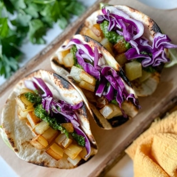 Three Spicy Potato Soft Tacos sit on a wooden cutting board surrounded by a yellow linen and parsley.
