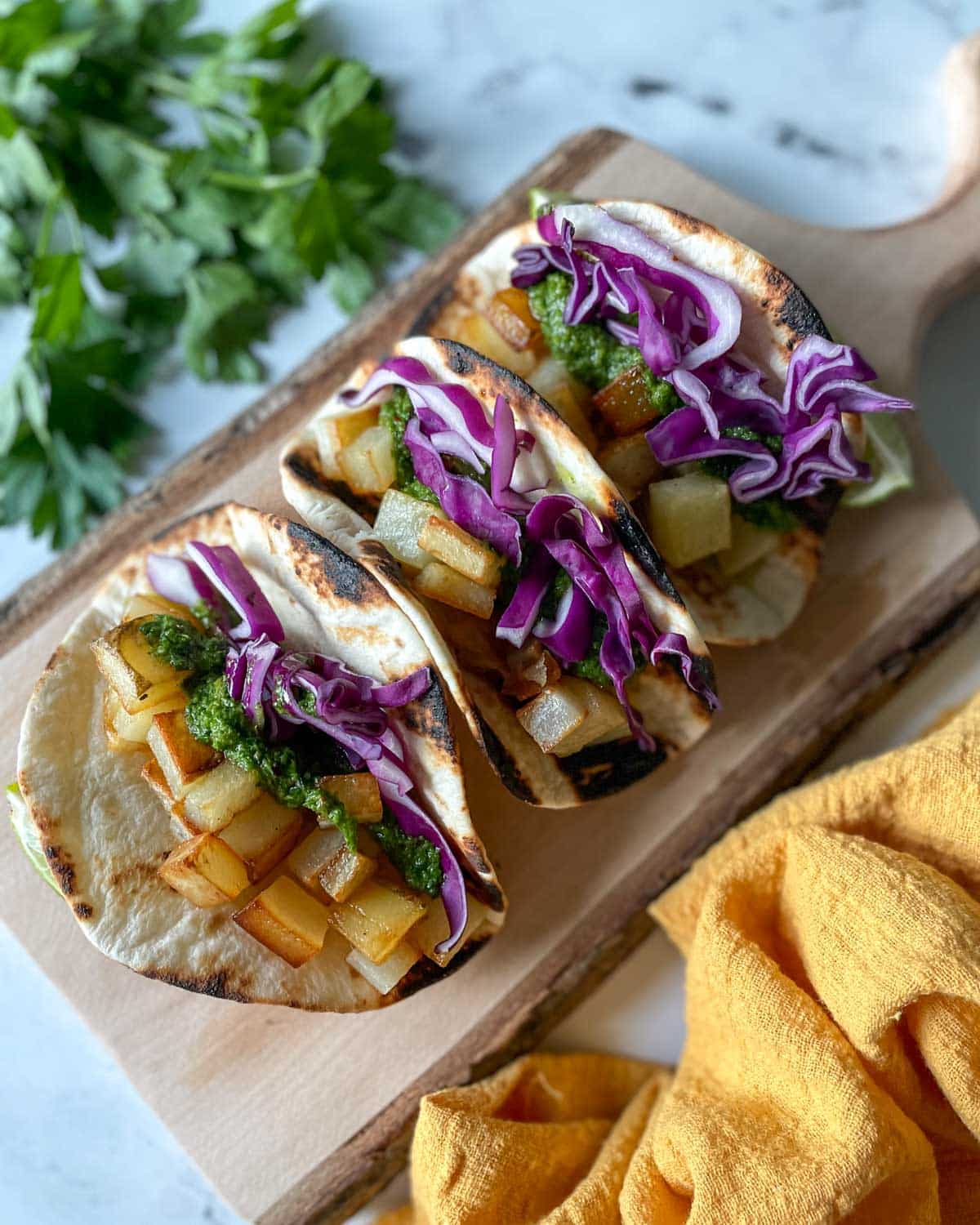 Three Spicy Potato Soft Tacos sit on a wooden cutting board surrounded by a yellow linen and parsley.