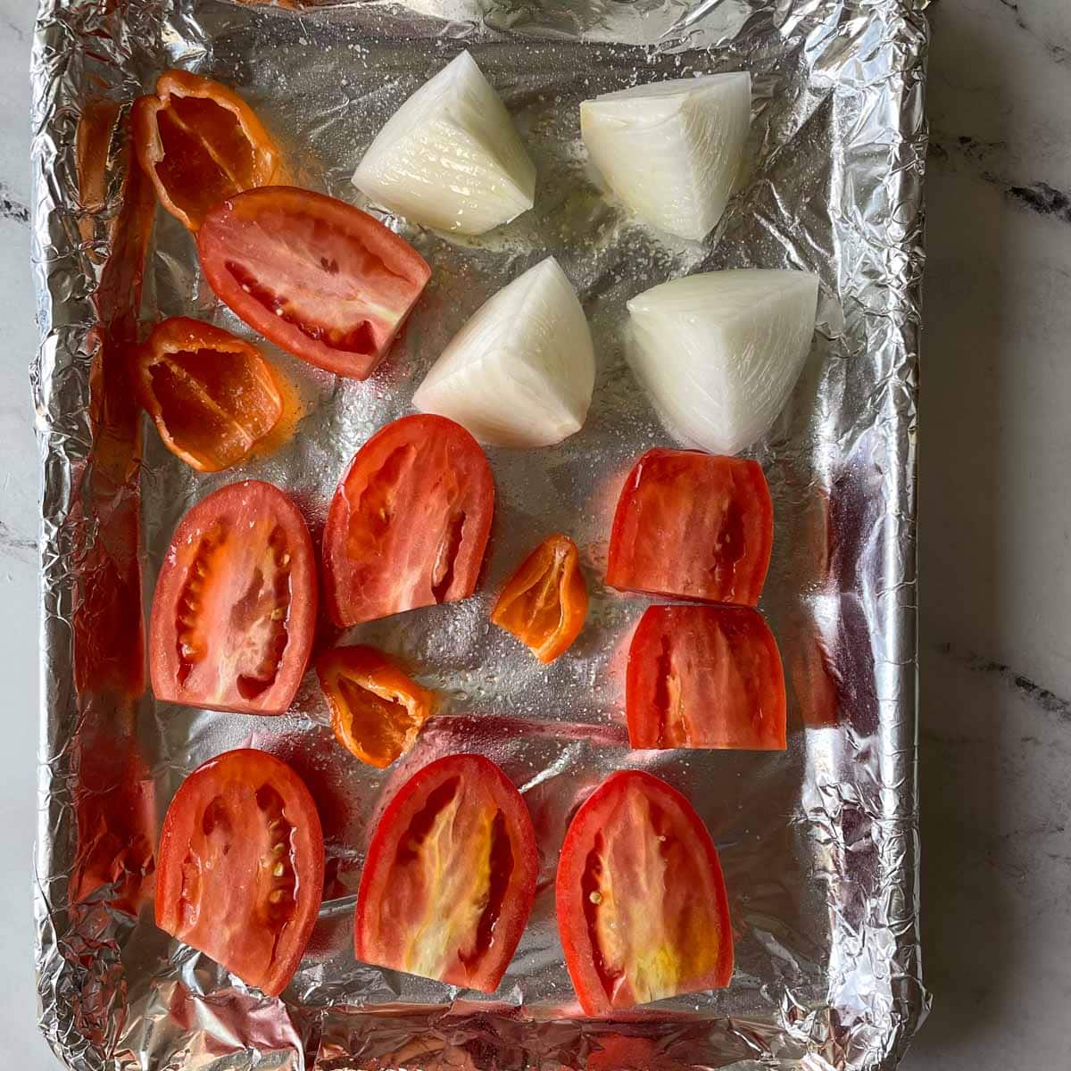 Halved tomatos and habaneros and half of a quartered white onion are shown a sheet tray covered with tin foil.