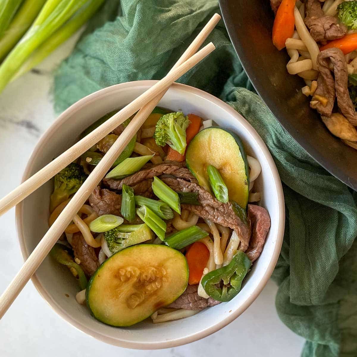 A bowl of beef yaki udon with chopsticks is shown beside a wok full of yaki udon, scallions, and a green linen.