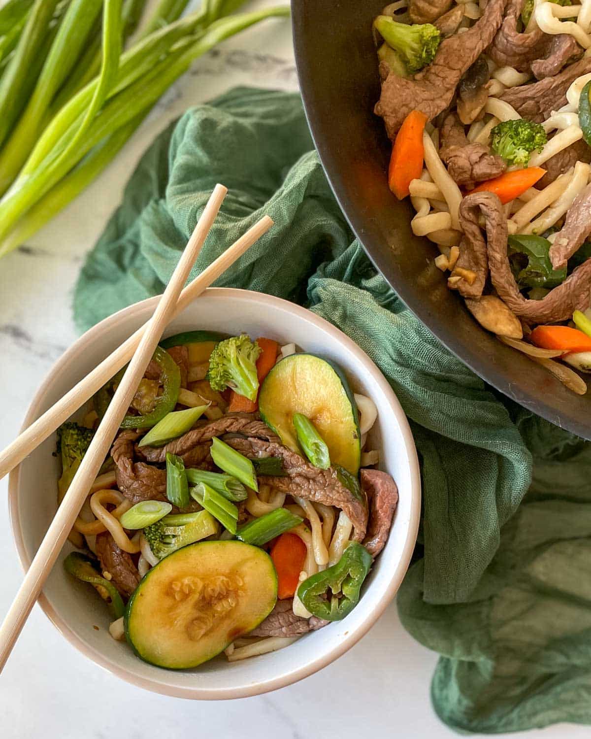 A bowl of beef yaki udon with chopsticks is shown beside a wok full of yaki udon, scallions, and a green linen.