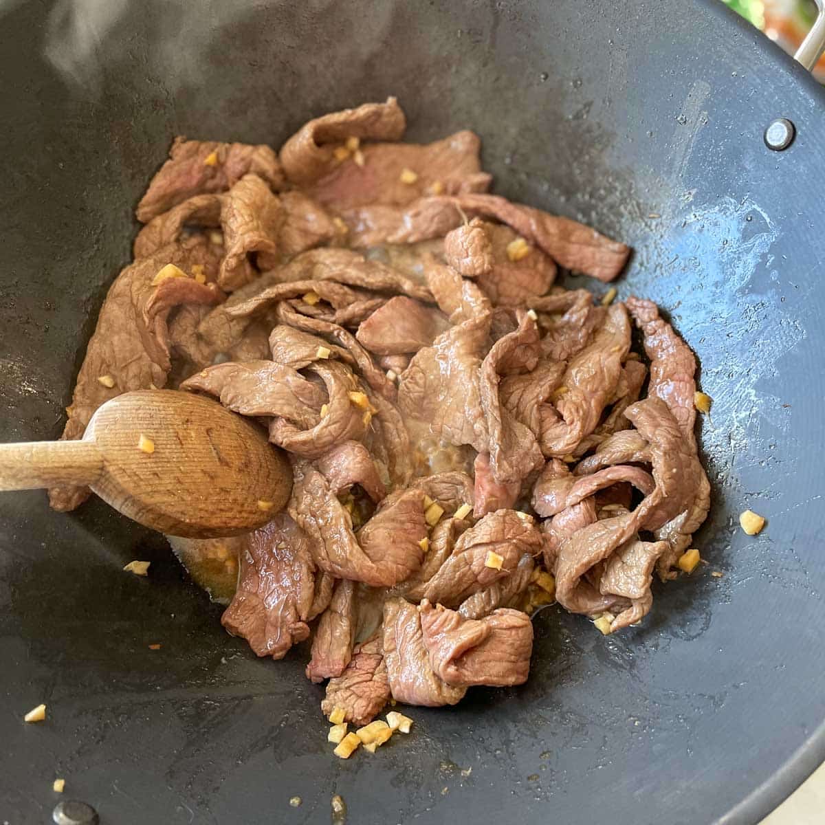 Thinly sliced beef is stirred in a black wok with a wooden spoon.