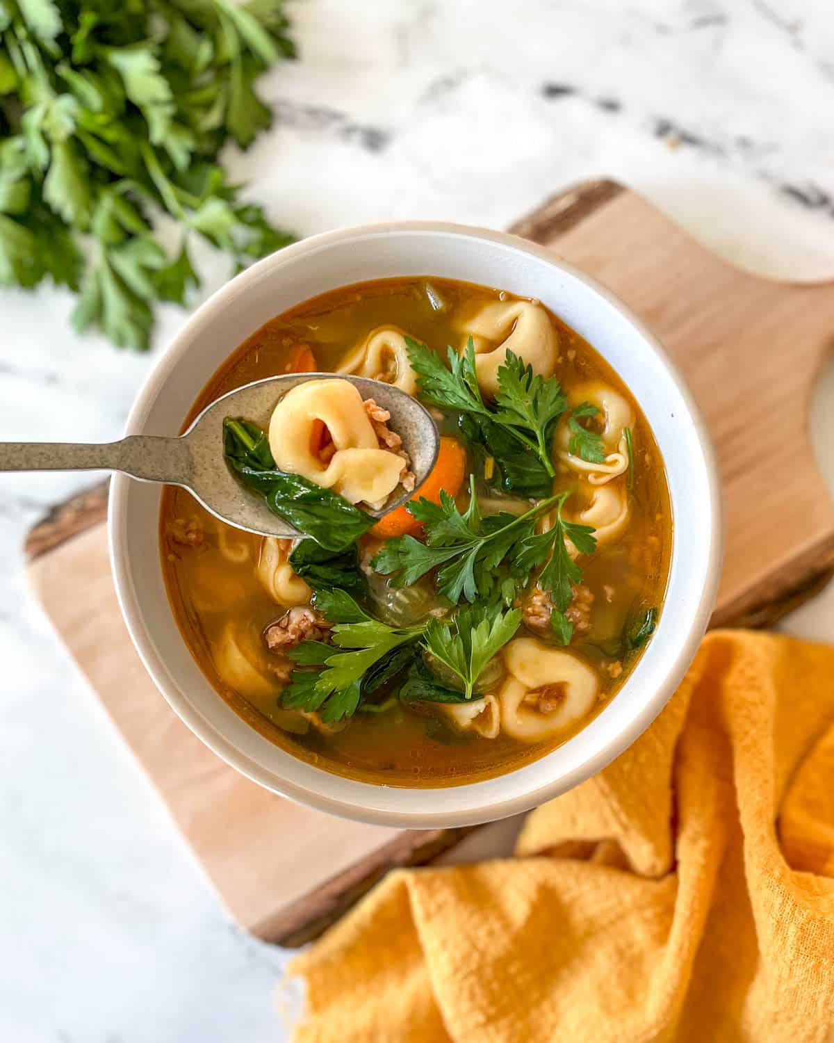 A bowl of tortellini soup sits on a rustic wooden cutting board with a yellow linen and a bunch of parsley.