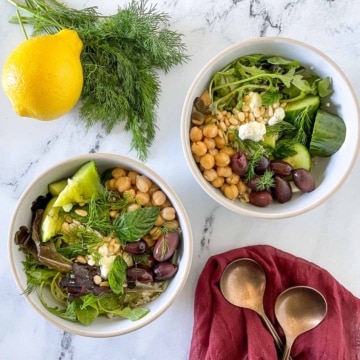 Two grain bowls are shown on a white marble counter with forks, a red linen, dill, and a lemon.