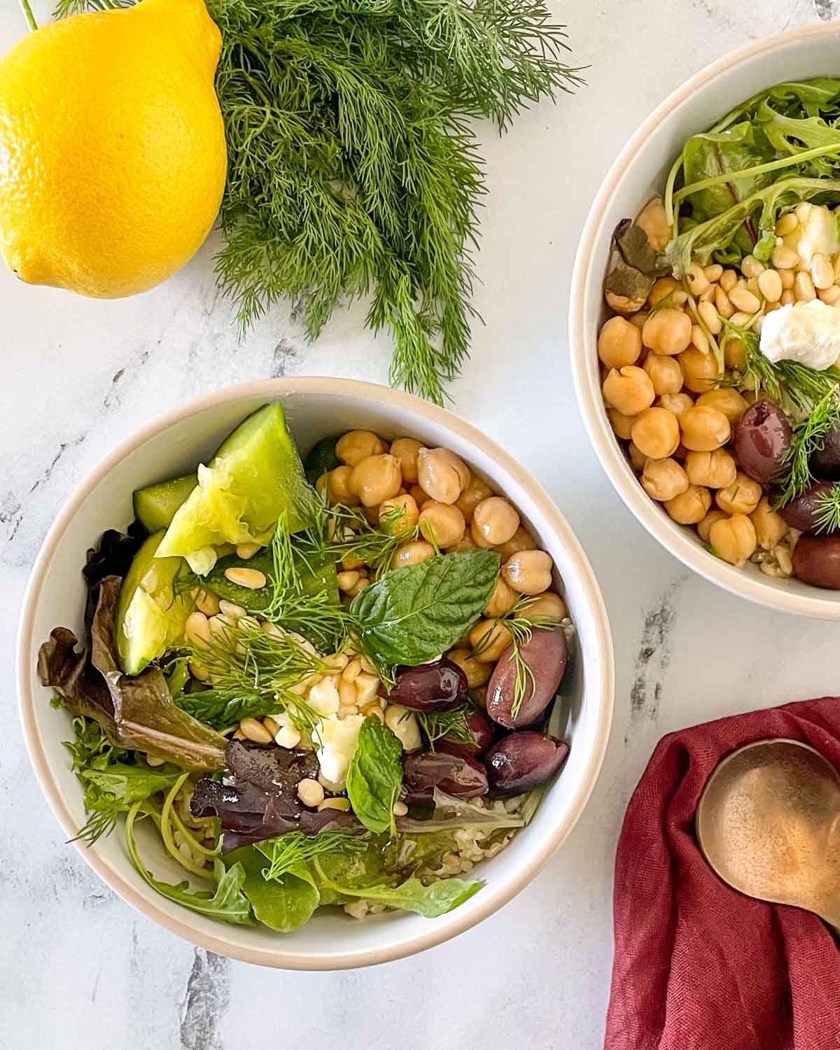 Two grain bowls are shown on a white marble counter with spoons, a red linen, dill, and a lemon.