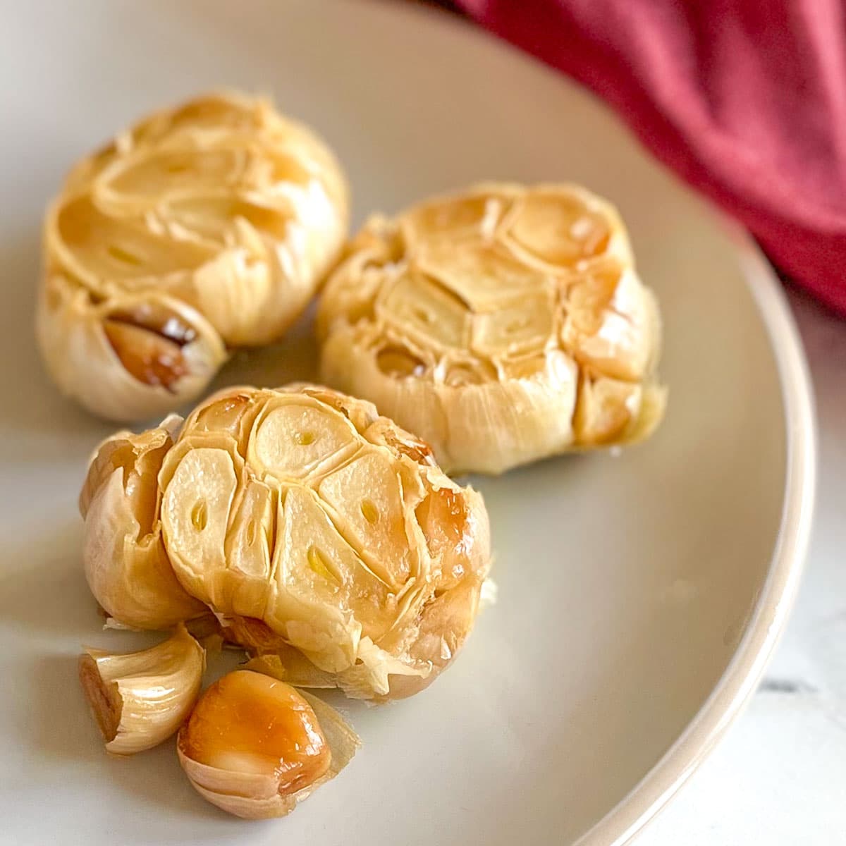 Three heads of roasted garlic sit on a white plate with a red linen in the background.