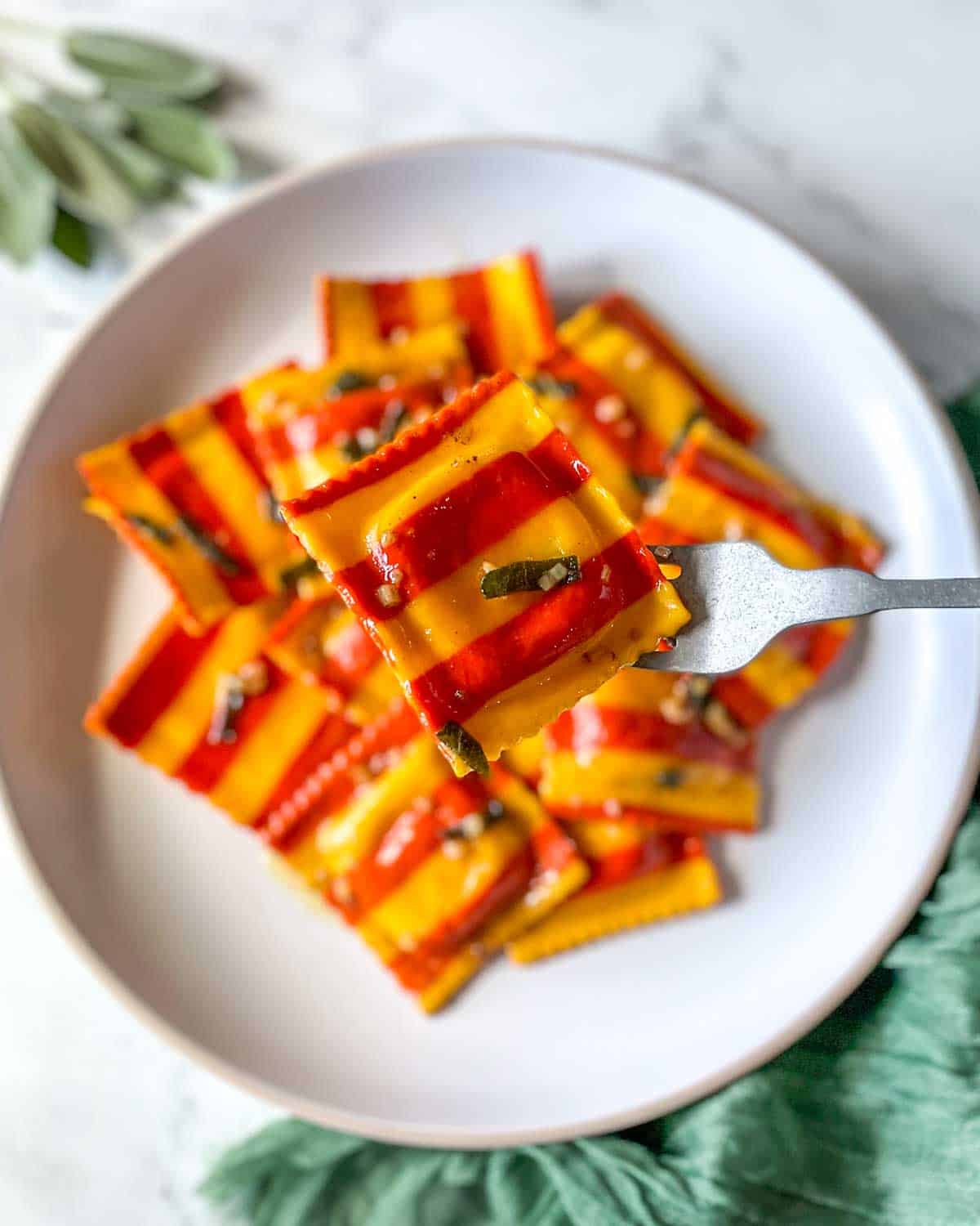 A red and yellow striped lobster ravioli is held on a silver fork over a plate of ravioli.