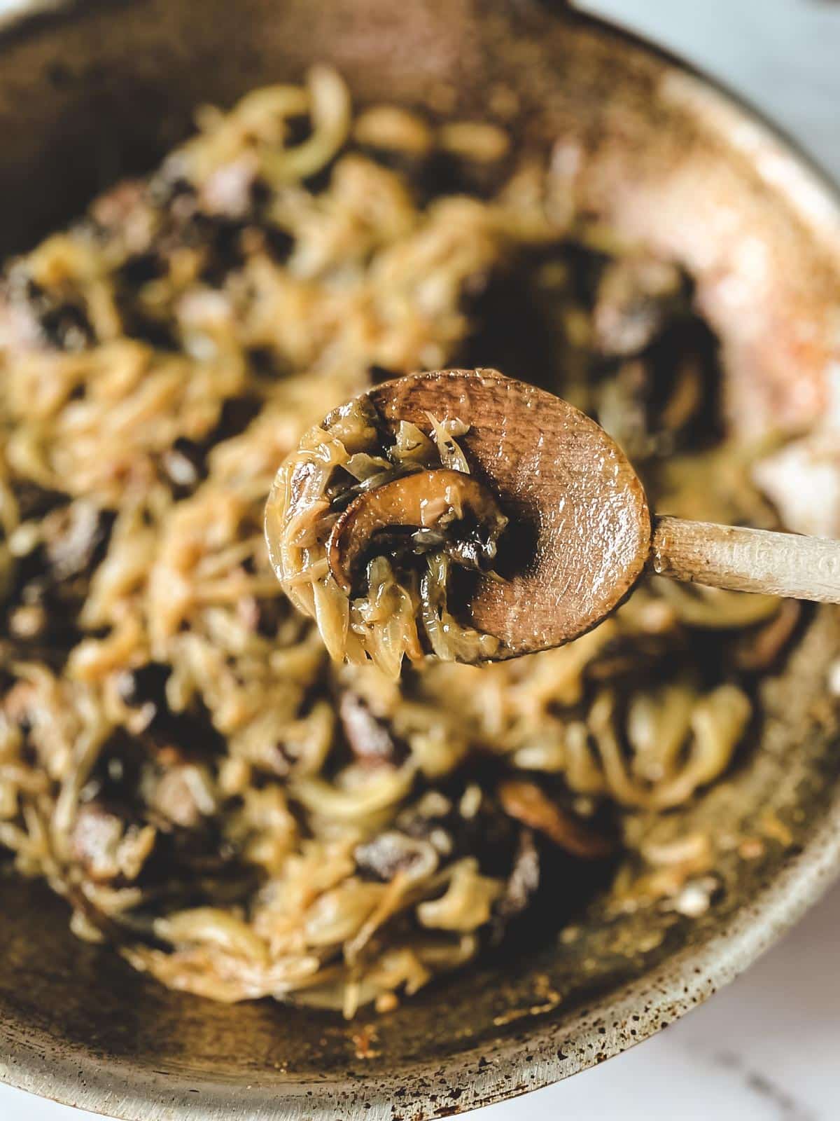 A wooden spoon offers a closeup view of caramelized mushrooms and onions above a pan in the background.