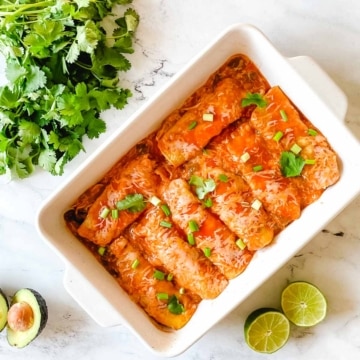 A baking dish full of chicken enchiladas is surrounded by cut limes, a halved avocado, and cilantro.