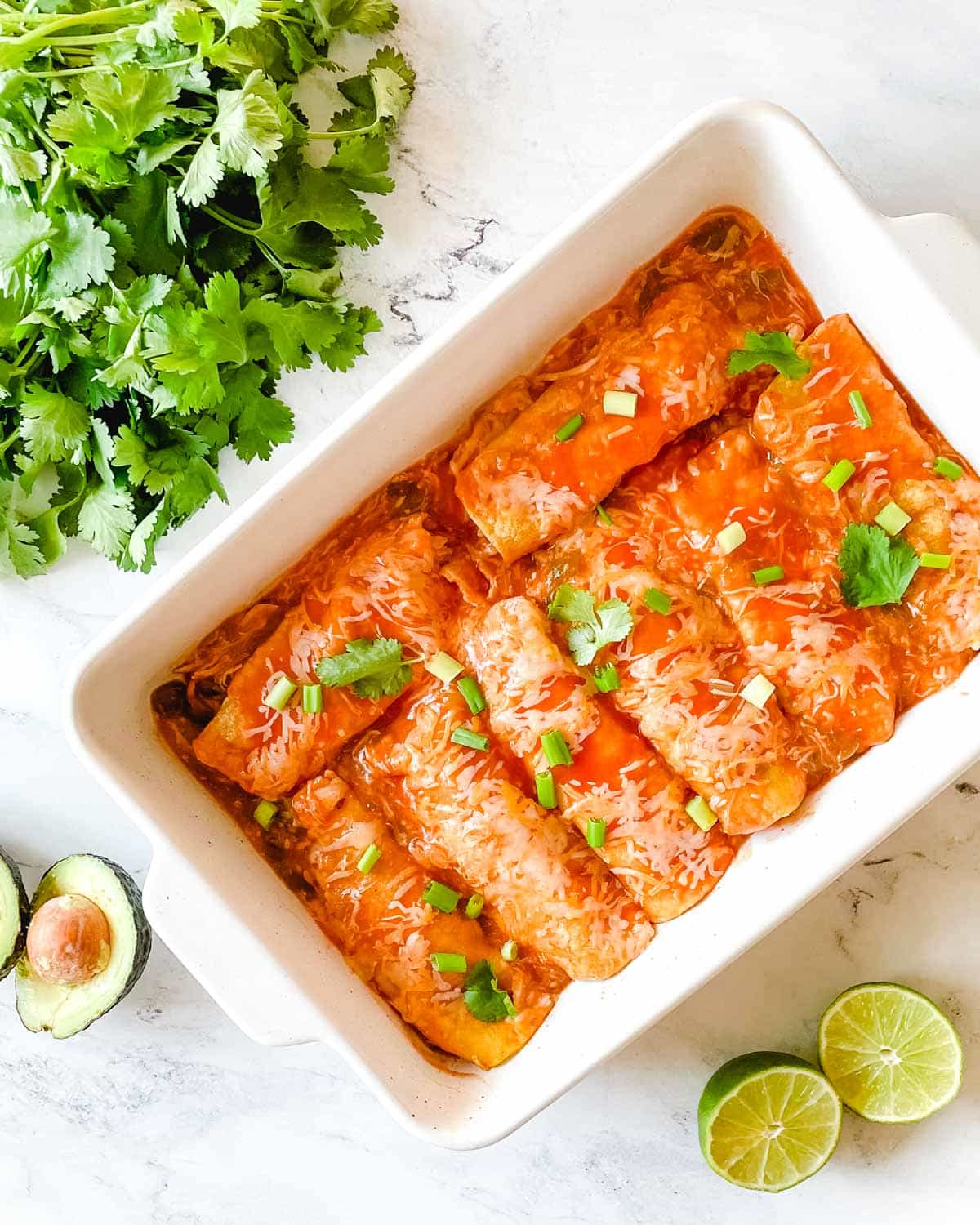 A baking dish full of chicken enchiladas is surrounded by cut limes, a halved avocado, and cilantro.
