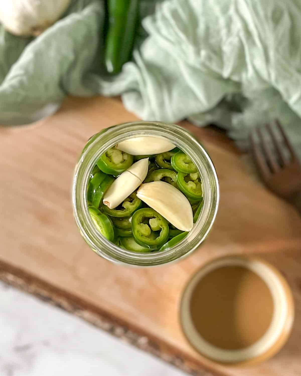 Pickled serrano peppers are shown in a glass jar surrounded by garlic, serrano peppers, and a fork.