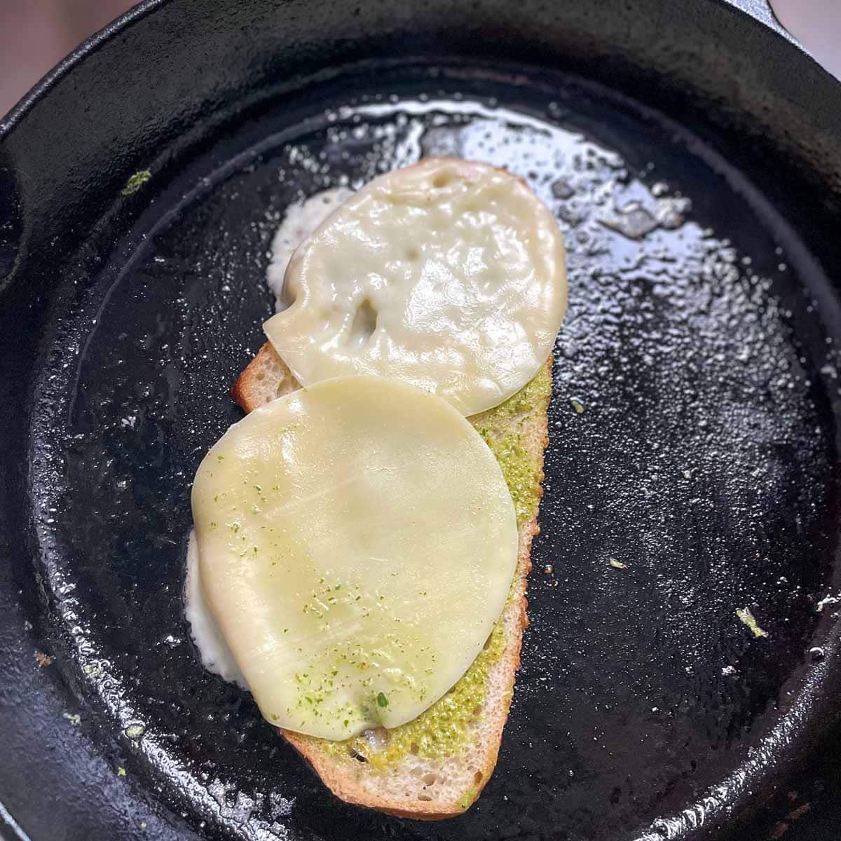 A slice of bread with cheese on top is shown toasting in a cast iron skillet.
