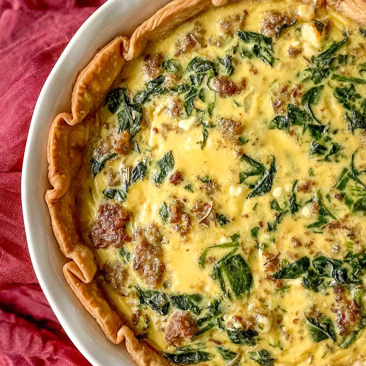 A closeup of a sausage spinach quiche is shown with a red linen.