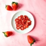 A white plate of air fryer dehydrated strawberries sits on a pink marble background surrounded by fresh strawberries.