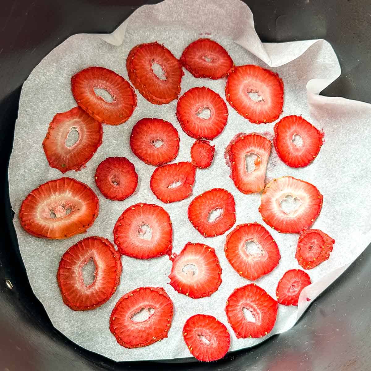 Partially dehydrated strawberry slices arranged in a single layer on parchment paper in an air fryer basket.