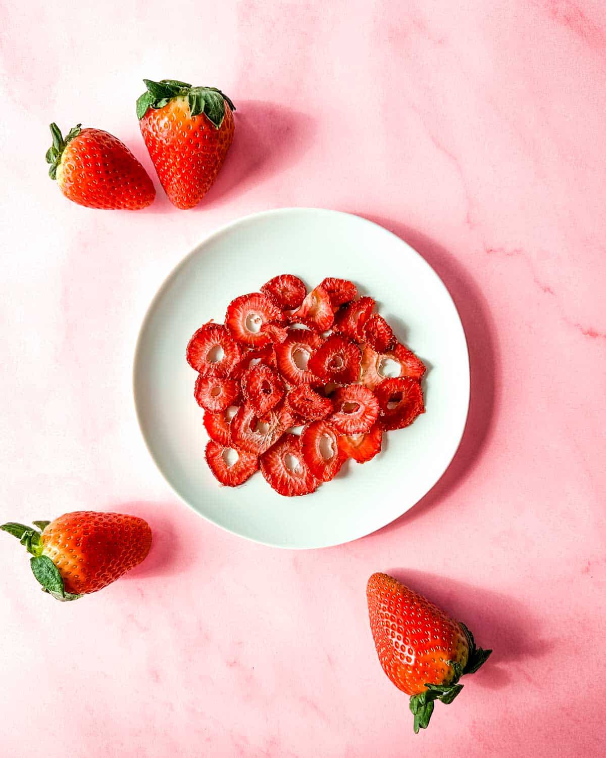 A white plate of air fryer dehydrated strawberries sits on a pink marble background surrounded by fresh strawberries.