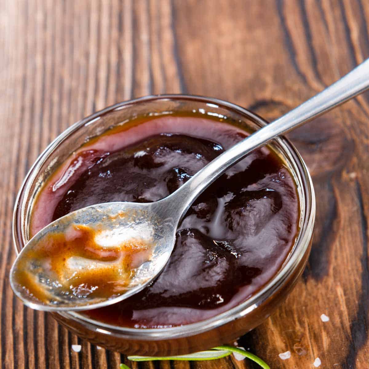 A dish of barbeque sauce sits on a wooden background with a spoon.