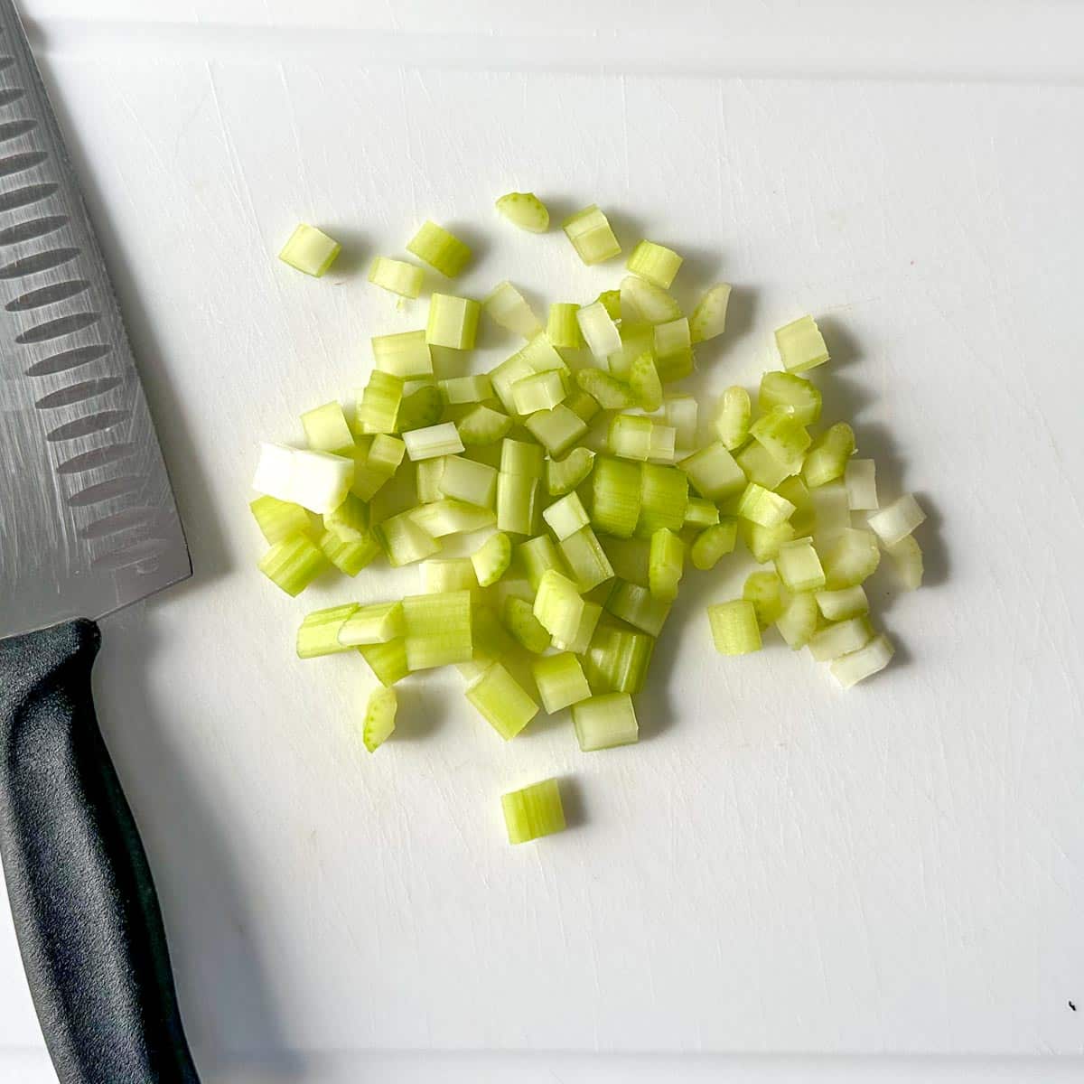 Chopped celery and a kitchen knife sit on a white cutting board.