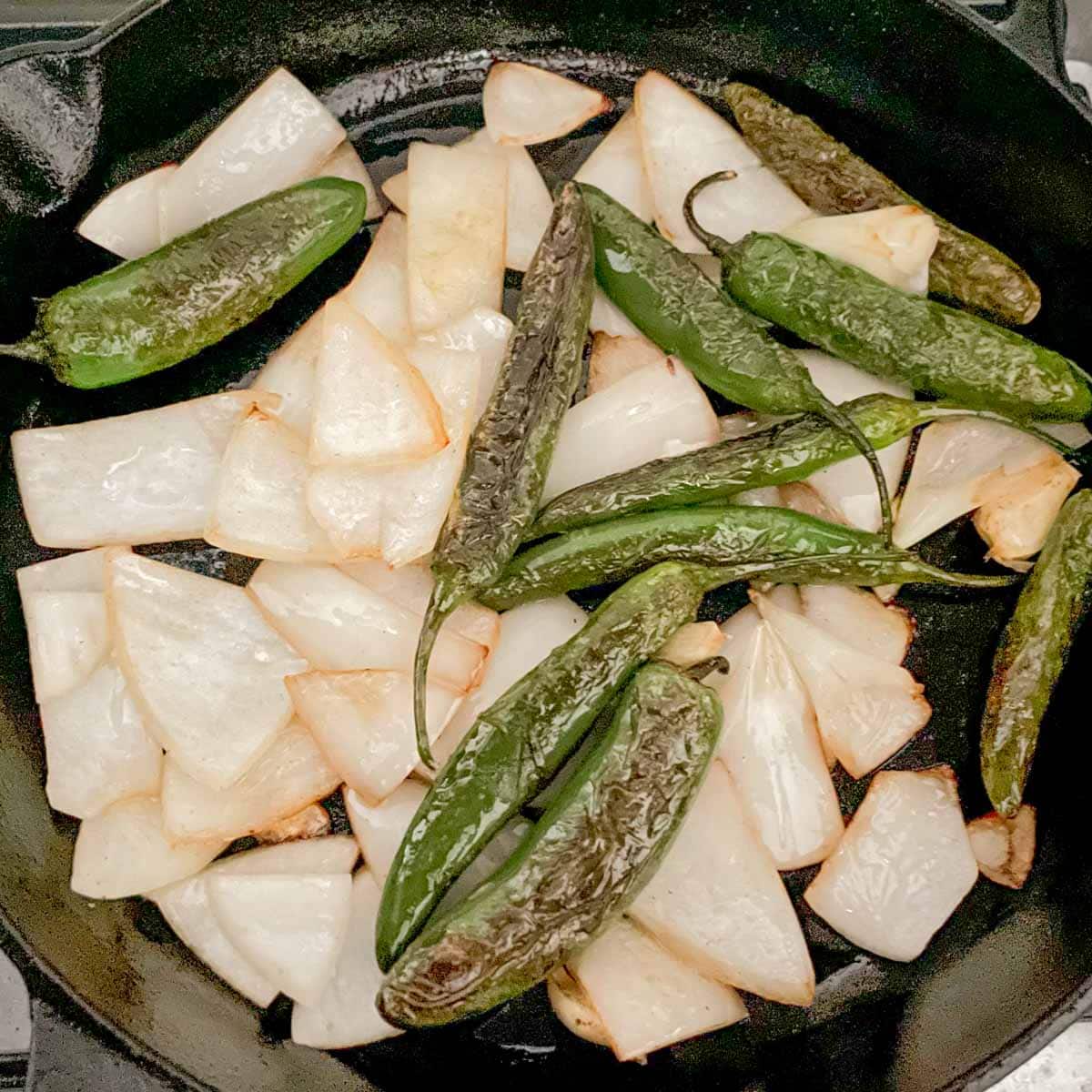 Cooked serranos, onion, and garlic are shown in a cast iron pan.