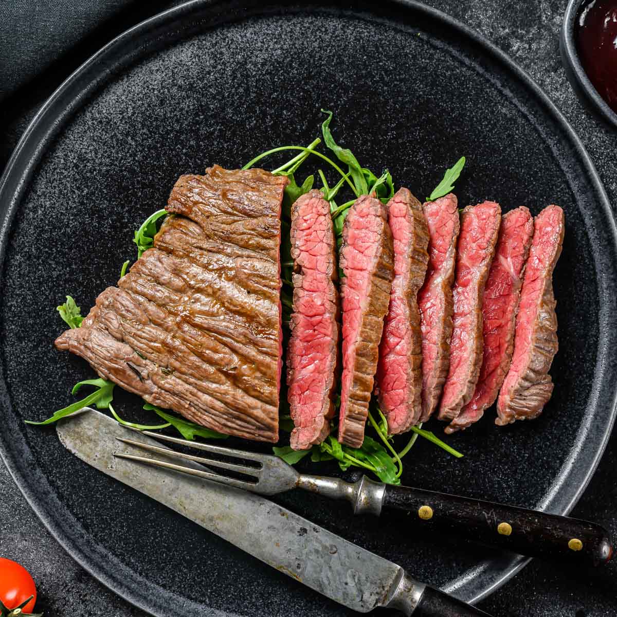 Cooked steak is shown on a black plate with black and silver fork and knife and arugula.