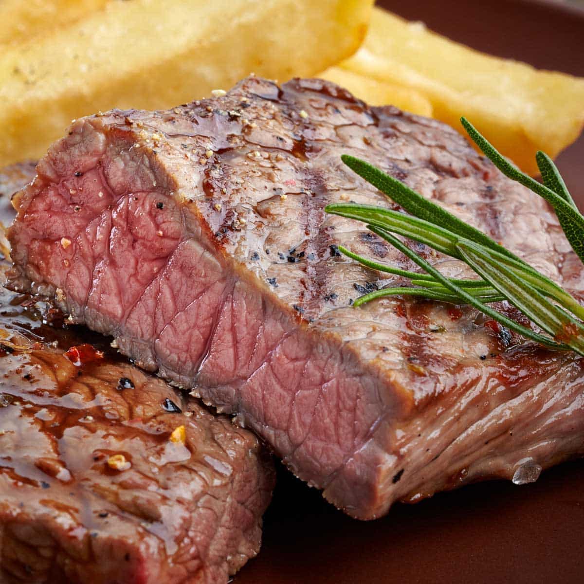A closeup is shown of a grilled steak sliced in half with a sprig of rosemary and fries in the background.