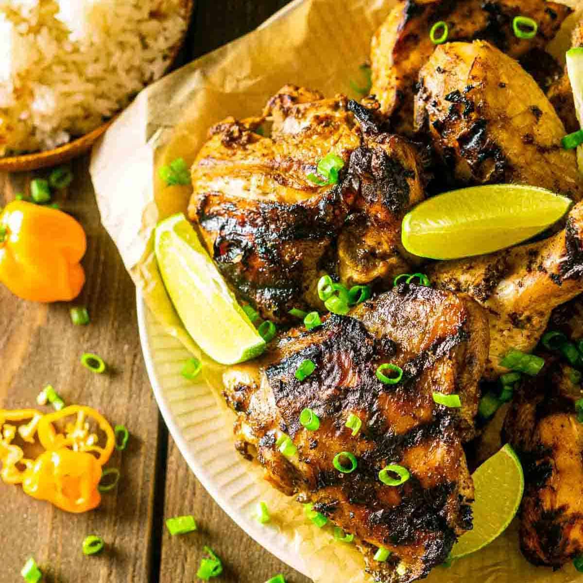 Jamaican jerk chicken sits on a white plate on a wooden table surrounded by sliced habaneros and green onions.