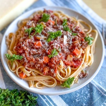 A bowl of linguine bolognese topped with parmesan cheese and parsely sits on a blue and white linen.