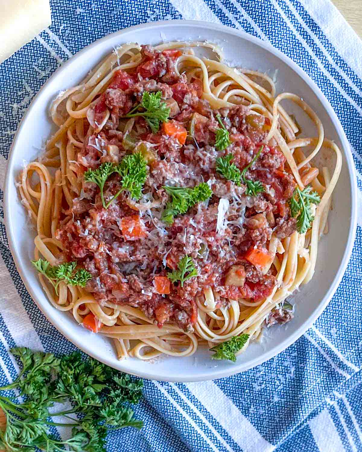 An overhead shot is shown of a dish of linguine bolognese topped with parmesan and parsley on a white and blue linen.