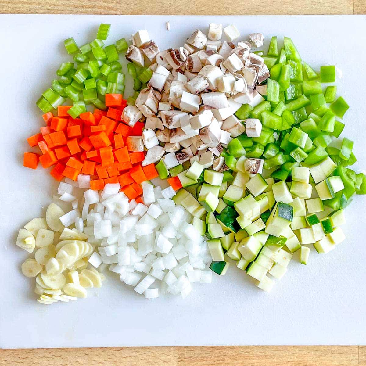 Diced carrot, celery, mushrooms, green bell pepper, zucchini, onion, and sliced garlic sit on a white cutting board.