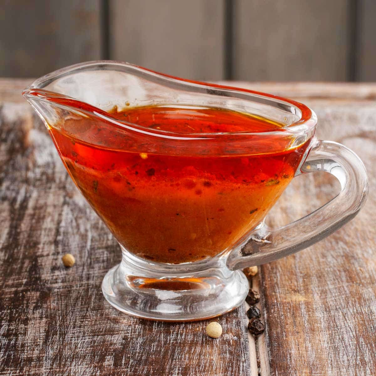 A glass gravy boat of red marinade is shown on a wooden table.