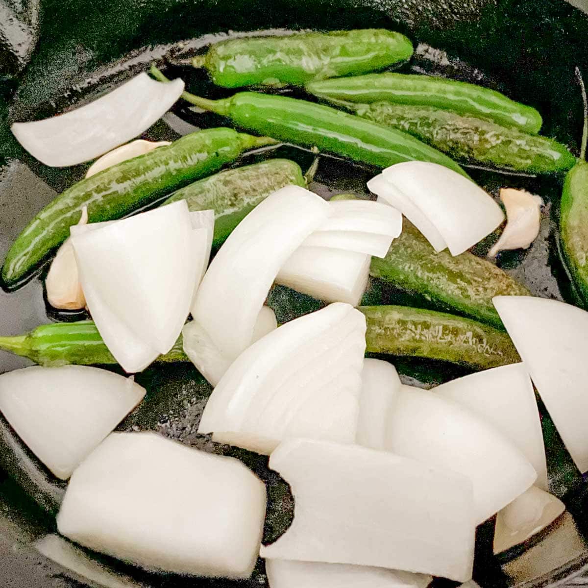 Serrano, onion, and garlic is shown roasting in a cast iron skillet.