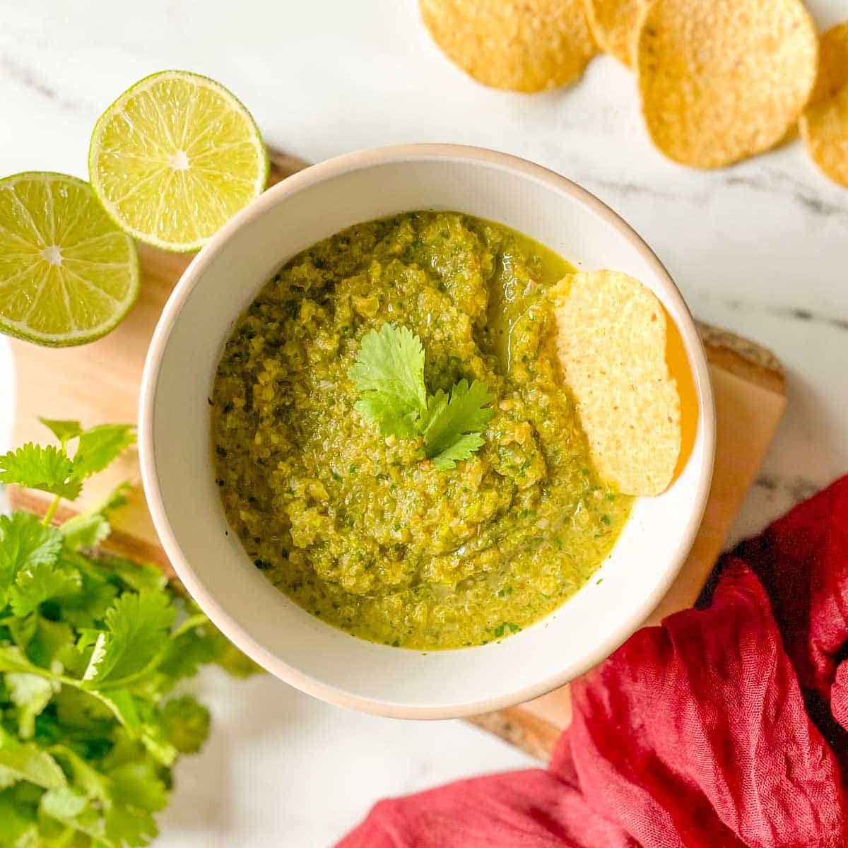 A bowl of serrano salsa is shown with limes, chips, cilantro, and a red linen.