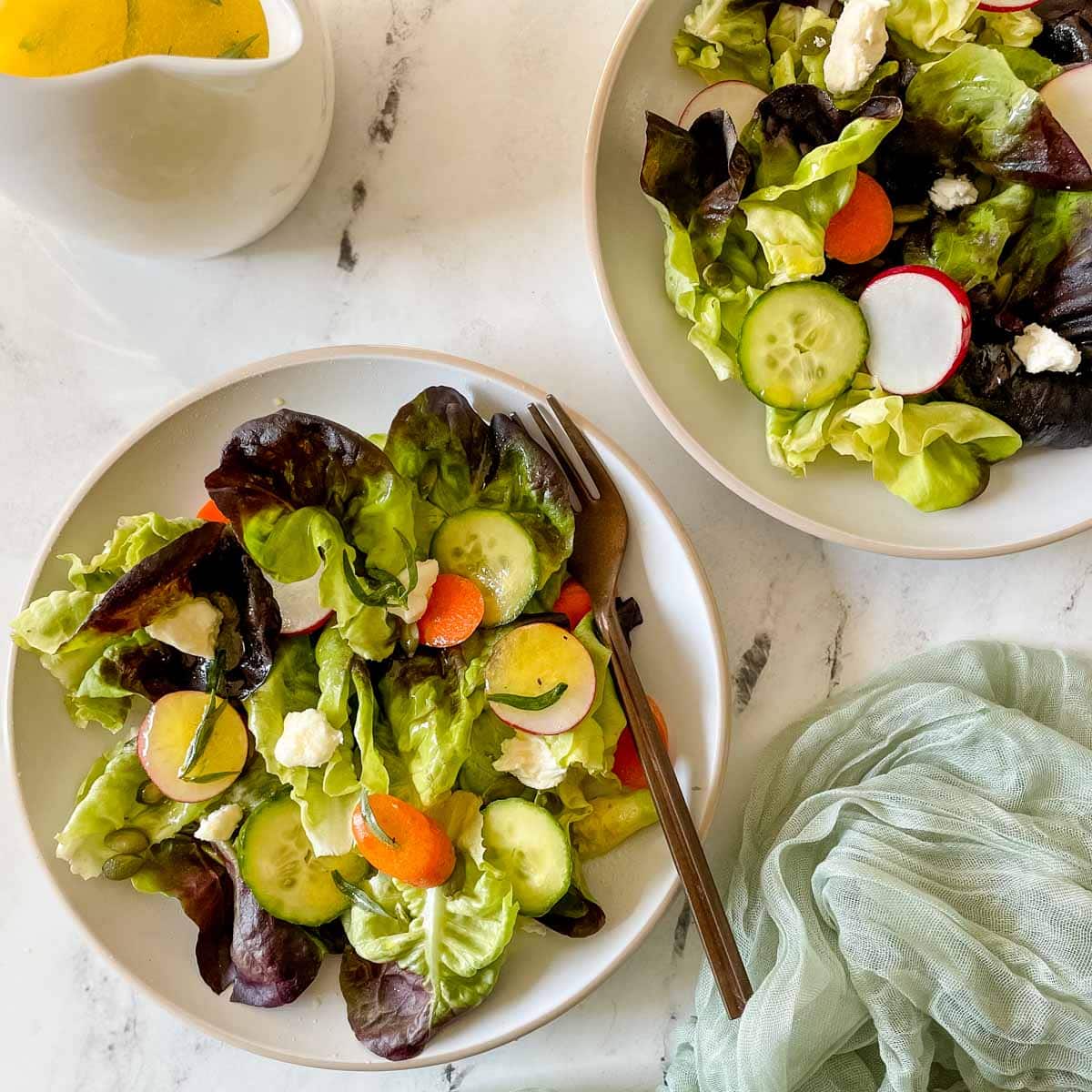 Two dishes of side salad are shown on a white marble counter with a small pitcher of lemon herb dressing and a green linen.