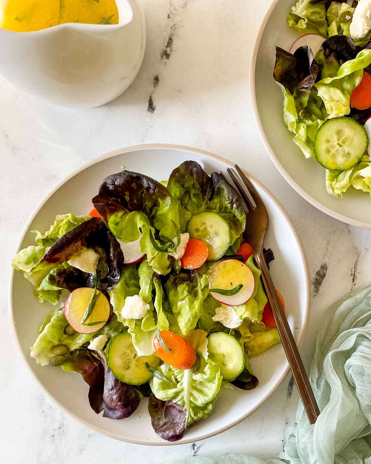 Two dishes of side salad are shown on a white marble counter with a small pitcher of lemon herb dressing and a green linen.