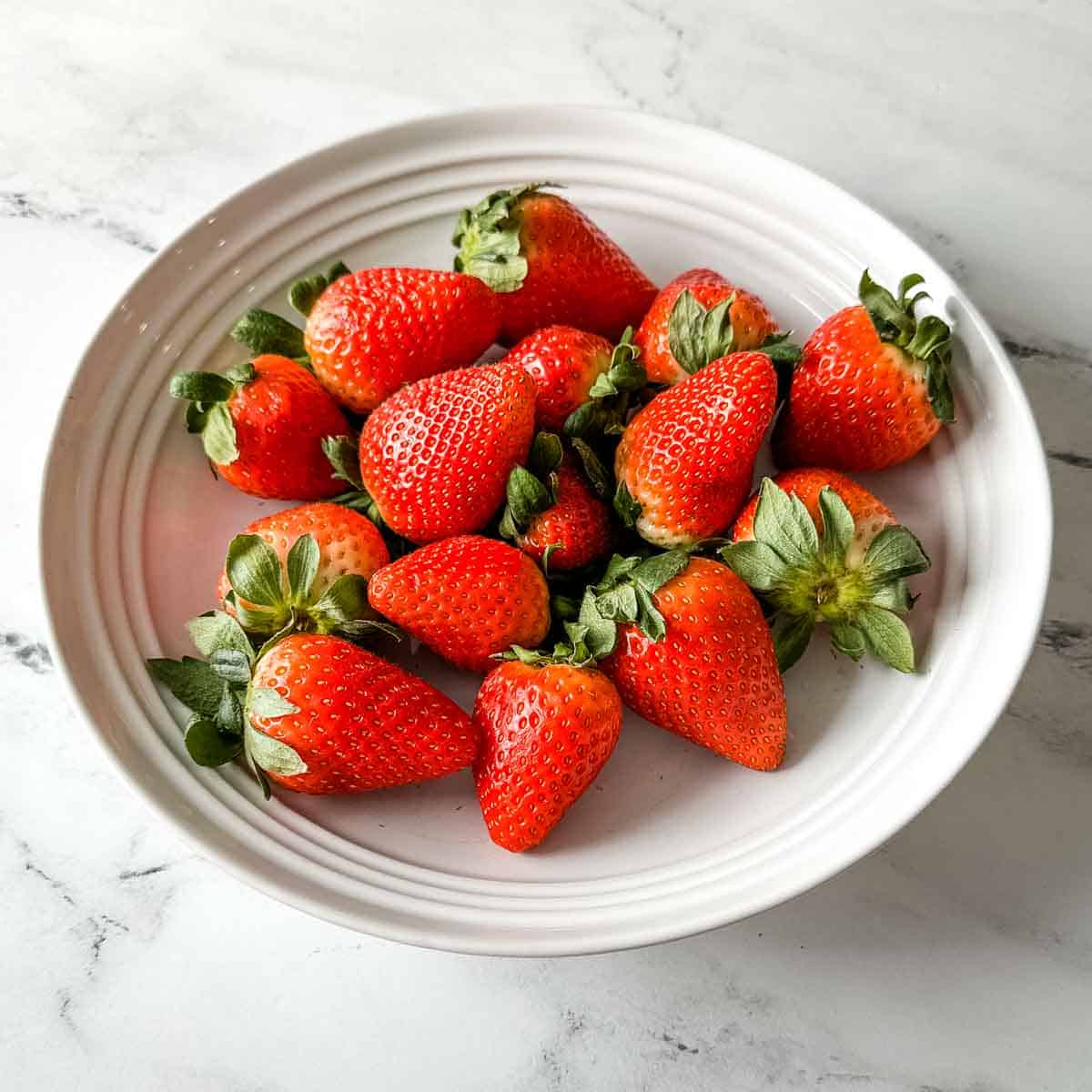 Fresh strawberries sit in a white bowl on a white marble counter.