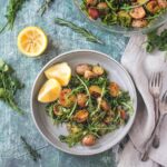 A bowl of arugula and roasted potato salad sits on a blue backdrop with lemons and herbs.