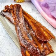 Glistening strips of candied bacon sit on a gray countertop with maple syrup in the background.