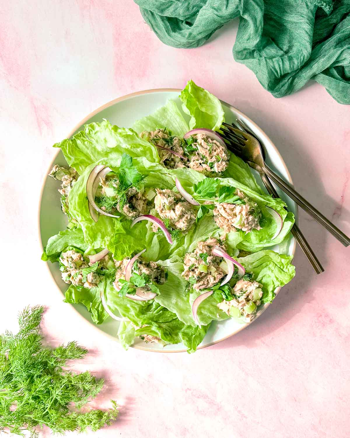 Healthy tuna salad without mayo sits on a bed of iceberg lettuce on a pink background surrounded by dill and a green linen.
