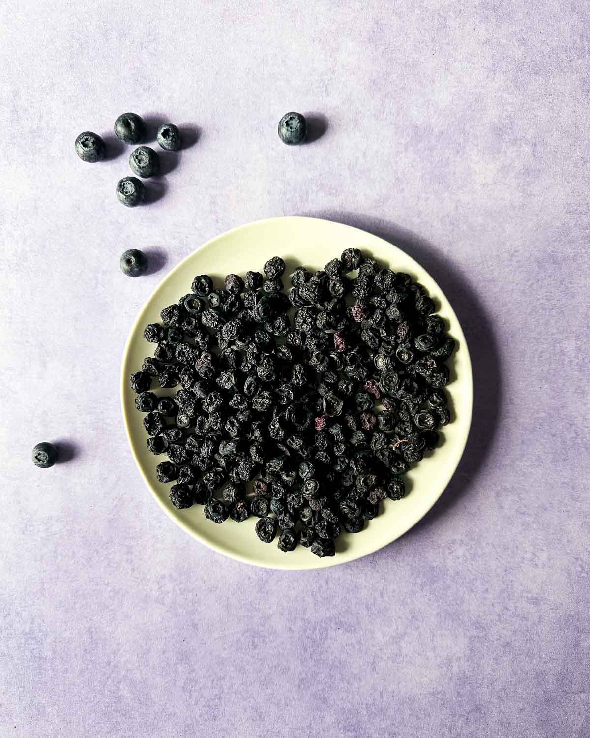 Air fryer dehydrated blueberries on a white plate on a purple backdrop surrounded by fresh blueberries.