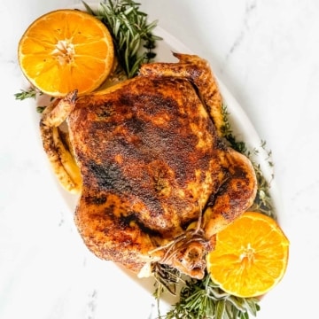 Christmas Roast Chicken on a white platter with halved oranges and fresh herbs.