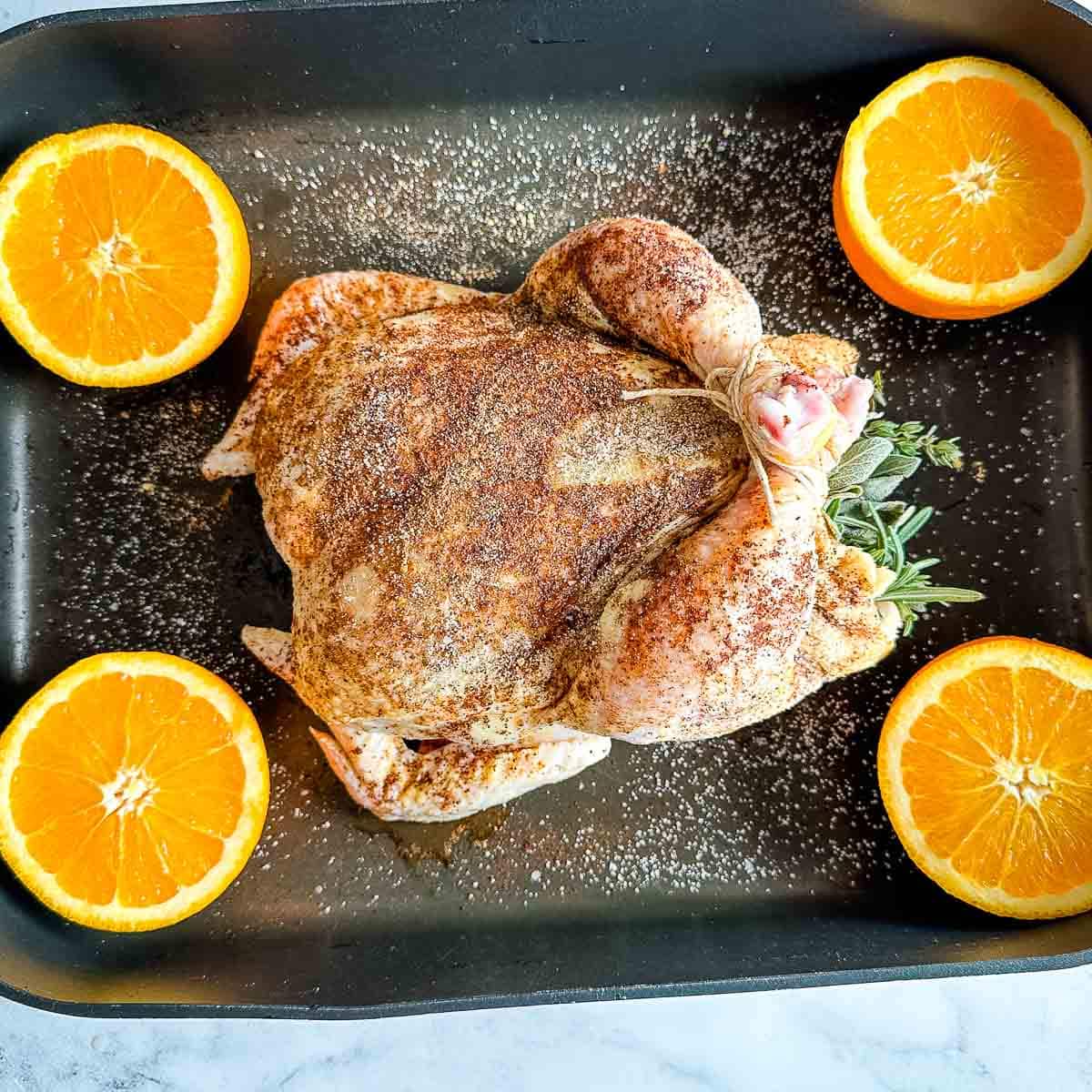 Raw Christmas Roast Chicken coated in spices and stuffed with herbs in a roasting pan with halved oranges.