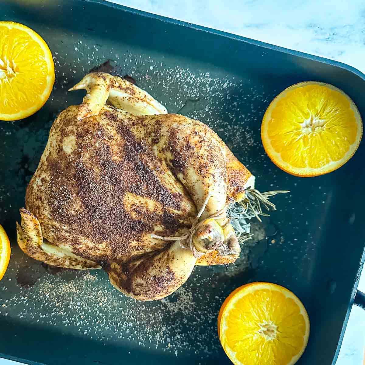 A half-cooked Christmas Roast Chicken in a roasting pan with halved oranges.