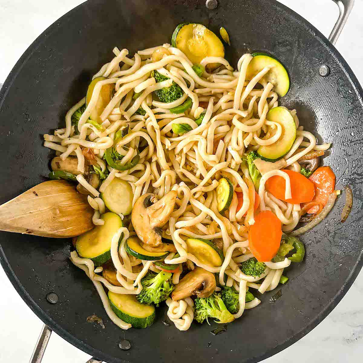 Udon noodles and mixed vegetables are stir-fried in a black wok with a wooden spoon.