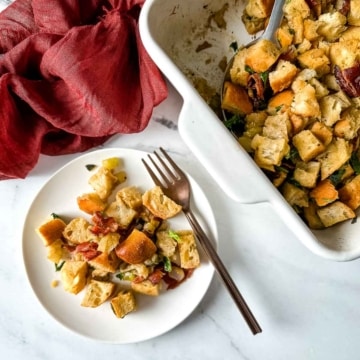 A baking dish of sage and bacon stuffing with a scoop taken out sits next to small plate of stuffing, a red linen, and a silver serving spoon.