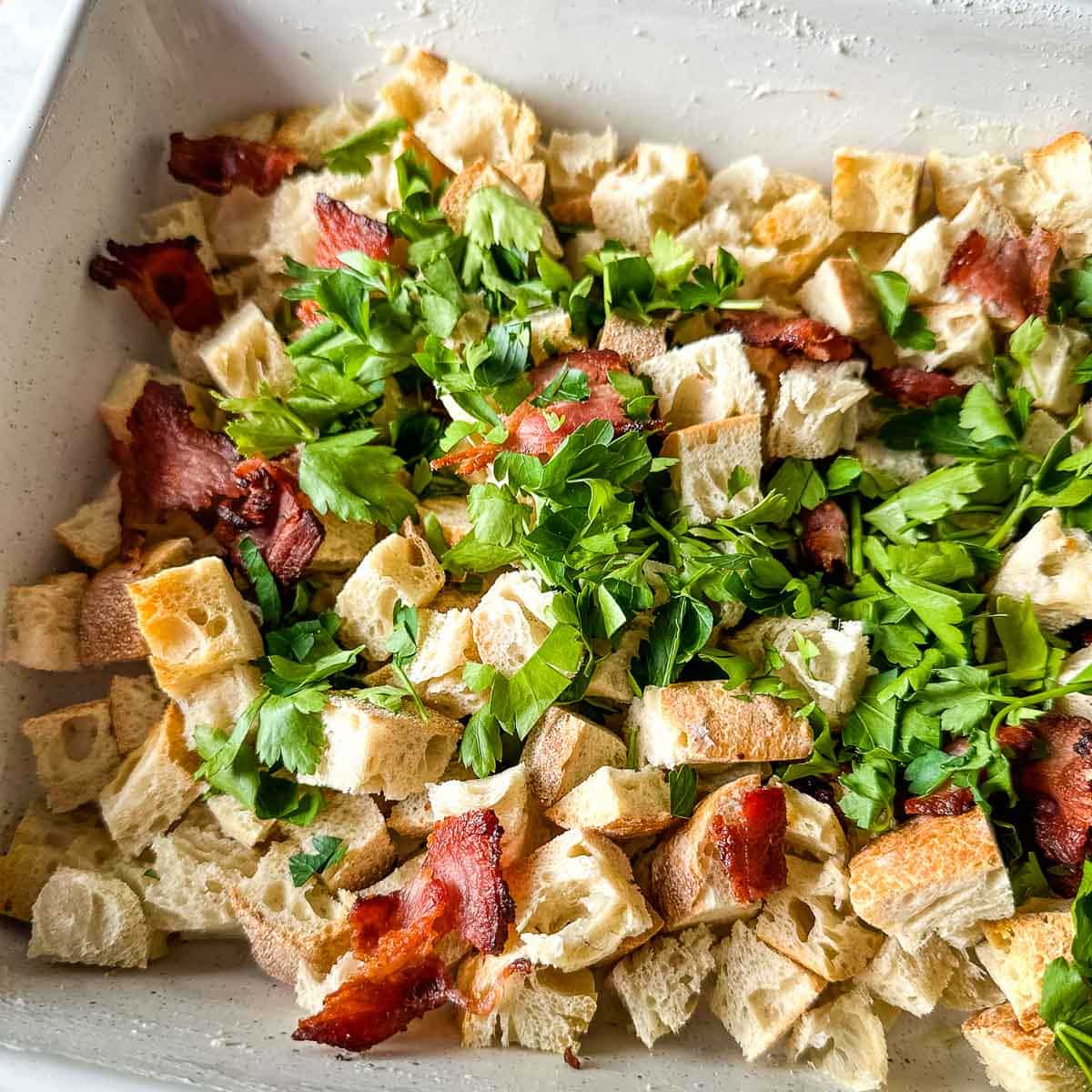 Bread cubes, parsley, and bacon sit in a buttered baking dish.