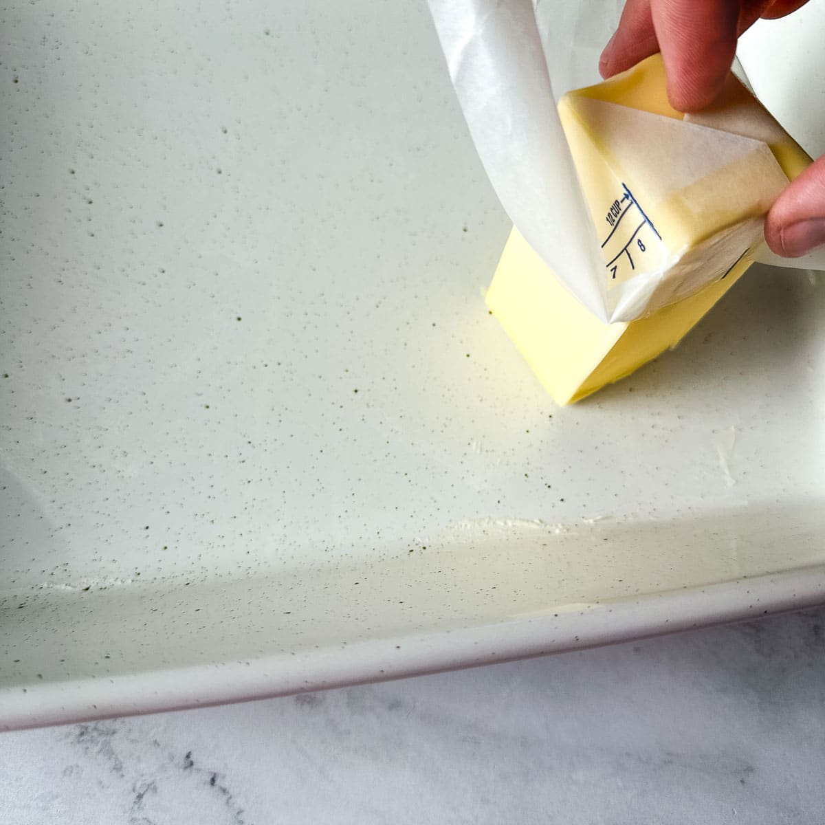 A stick of butter is rubbed onto a white baking dish.
